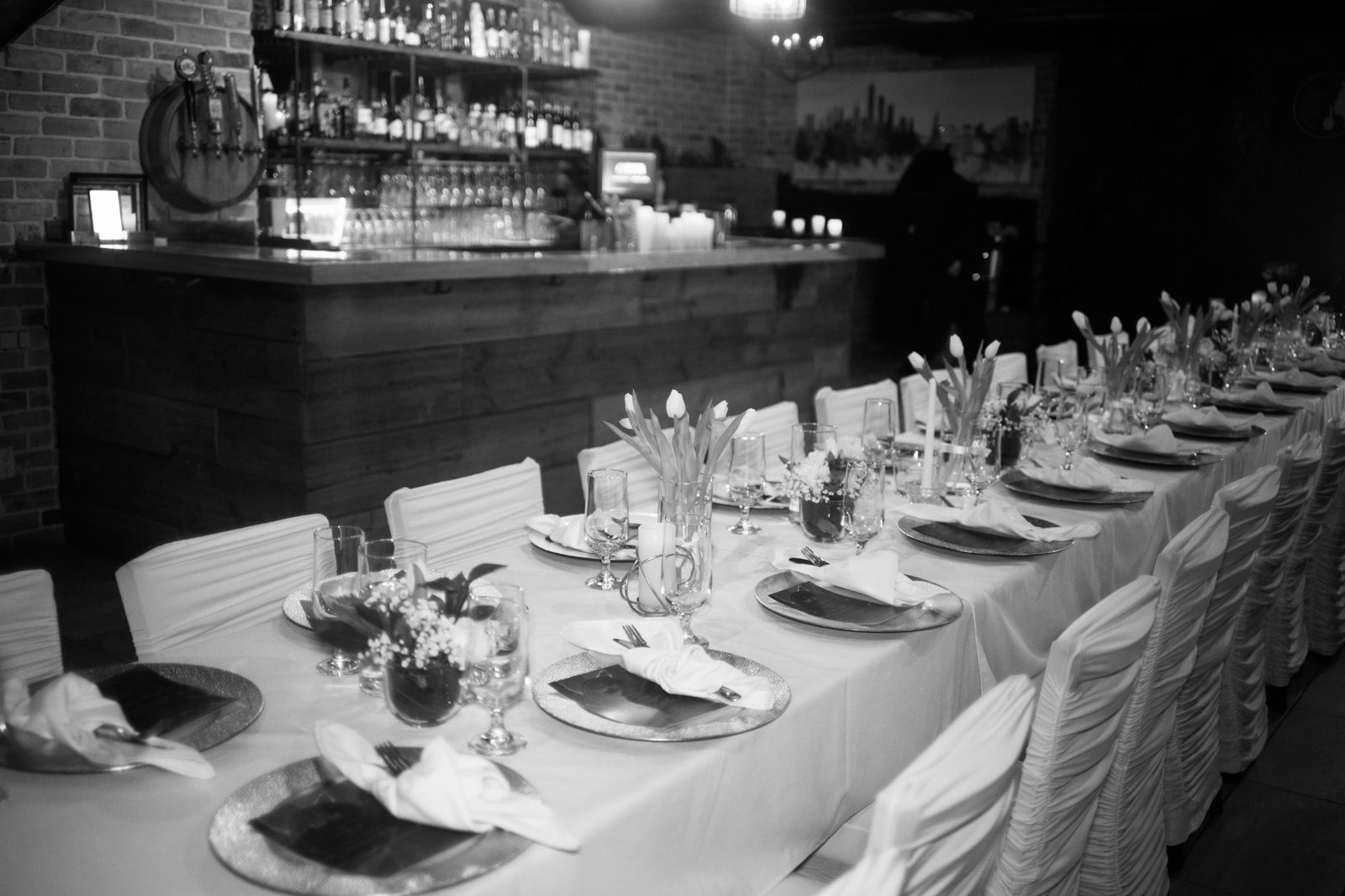 One long table laid for a seated dinner in front of the bar, set with tulips, chargers and folded napkins